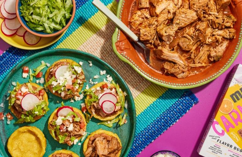 Overhead of mini tostadas, shredded meat, and colorful packaging on a woven mat.